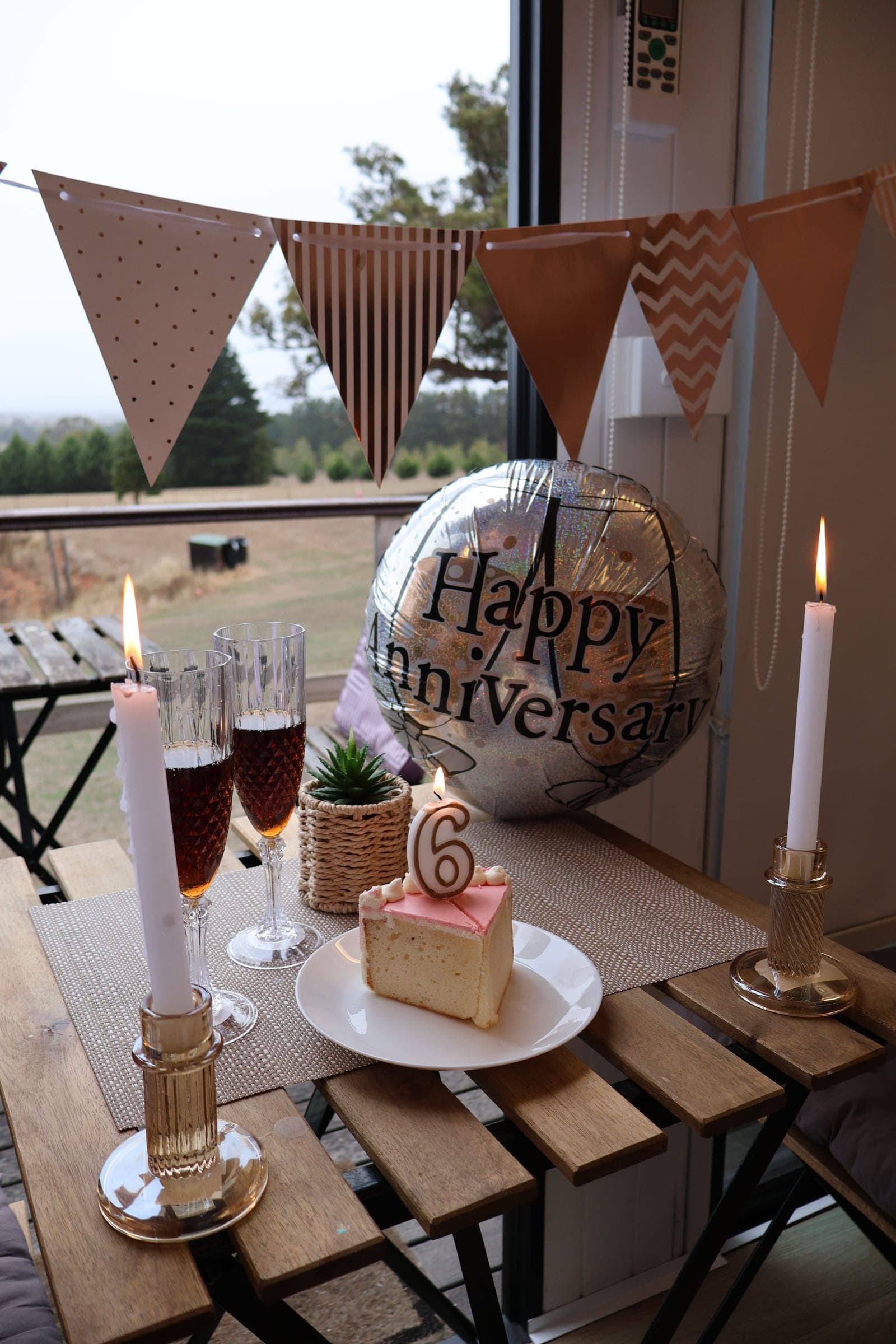A table laid for an anniversary — two glasses, lit candles, a slice of cake and a balloon