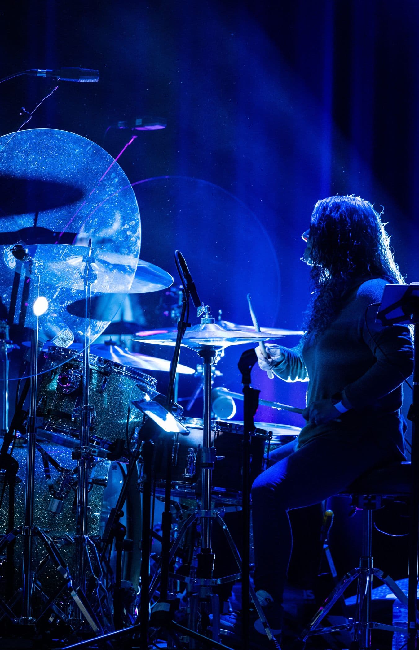 A drummer mid-beat at a glitter-finish kit, lit deep blue against a darkened stage