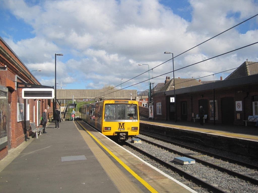 A yellow and white Tyne and Wear Metro train waiting at a suburban platform under a bright, cloudy sky